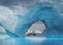 A small Zodiac inflatable boat carries tourists beneath a huge blue iceberg in the lagoons and bays surrounding the Antarctic peninsular