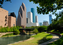 Modern skyscrapers of Houston, Texas are seen in this horizontal color image across a waterway flowing through a city park. A perfect, clear blue sky is a backdrop to this afternoon cityscape. Tops of the buildings are somewhat outlined by tree branches. 